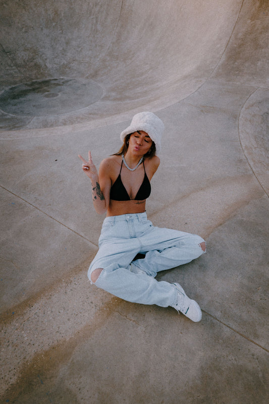 Marleen sitting casually in a skatepark bowl, wearing a black bikini top Sheila, ripped light jeans, white bucket hat, and sneakers, posing with a peace sign and playful expression. Peachys Wear Owner and Master Director.