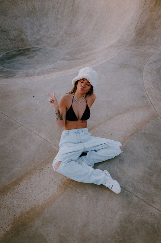 Marleen sitting casually in a skatepark bowl, wearing a black bikini top Sheila, ripped light jeans, white bucket hat, and sneakers, posing with a peace sign and playful expression. Peachys Wear Owner and Master Director.